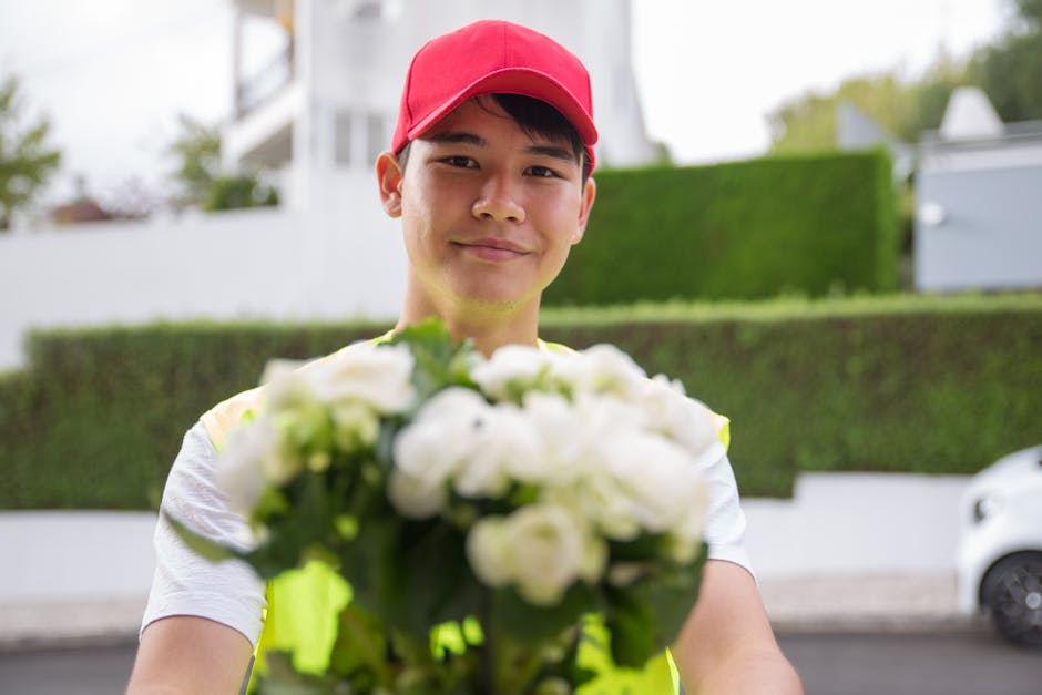 A delivery person wearing a red cap and a high-visibility vest is holding a bouquet of fresh white flowers, which include roses and other blooms, wrapped in clear cellophane and tied with a ribbon. Th