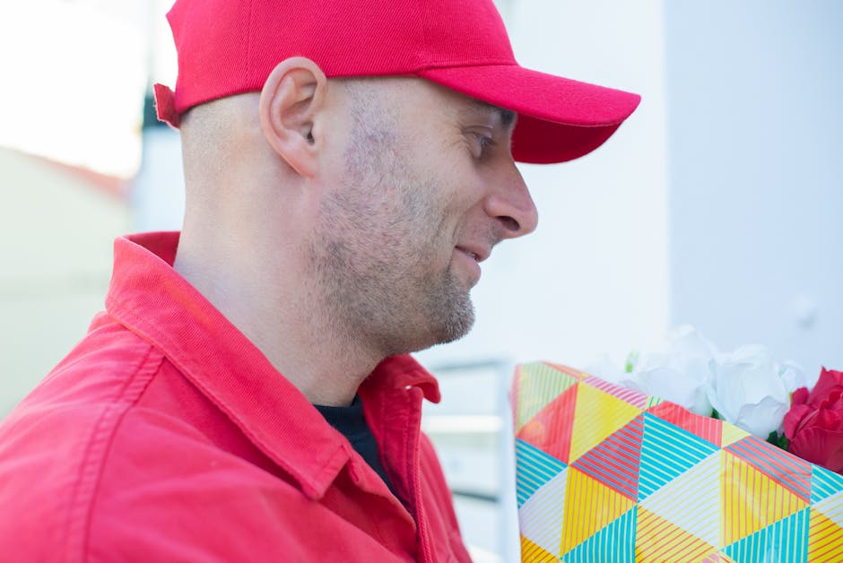 A delivery person dressed in a red cap and matching red jacket, holding a floral gift, which is wrapped in colorful geometric patterned paper with yellow, blue, red, and white shades, containing white