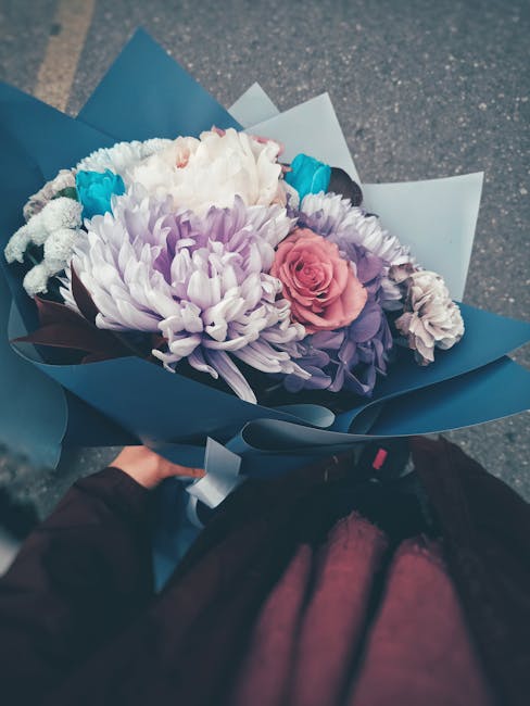 A bouquet of fresh flowers featuring a large, white chrysanthemum, pink roses, and purple lisianthus, arranged with dark green foliage. The flowers are wrapped in elegant blue and white paper with a d