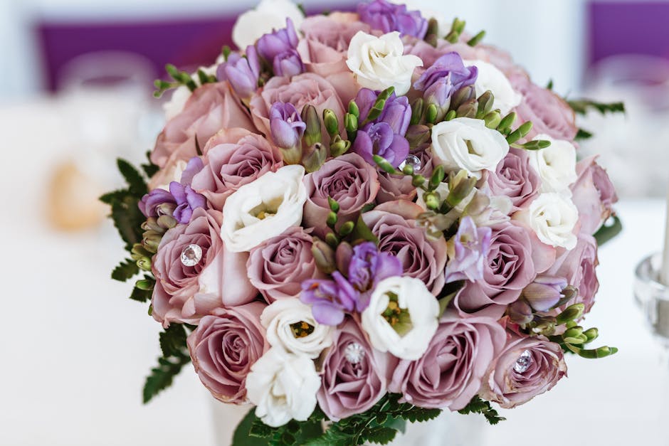 A close-up of a floral arrangement featuring a bouquet of fresh, pastel pink roses, white lisianthus, and purple freesias, all with vibrant green foliage. The bouquet is arranged in a rounded, compact